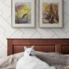 En interiörbild med två tavlor på väggen och en katt i sängen. Tavlorna är "Världarna vaknar av andetagen" och "Vilar i bladens dallrande dager". Två akvarellmålningar av Helene Engström.
