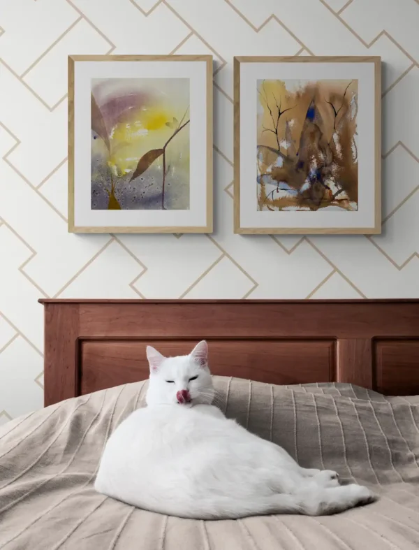 En interiörbild med två tavlor på väggen och en katt i sängen. Tavlorna är "Världarna vaknar av andetagen" och "Vilar i bladens dallrande dager". Två akvarellmålningar av Helene Engström.